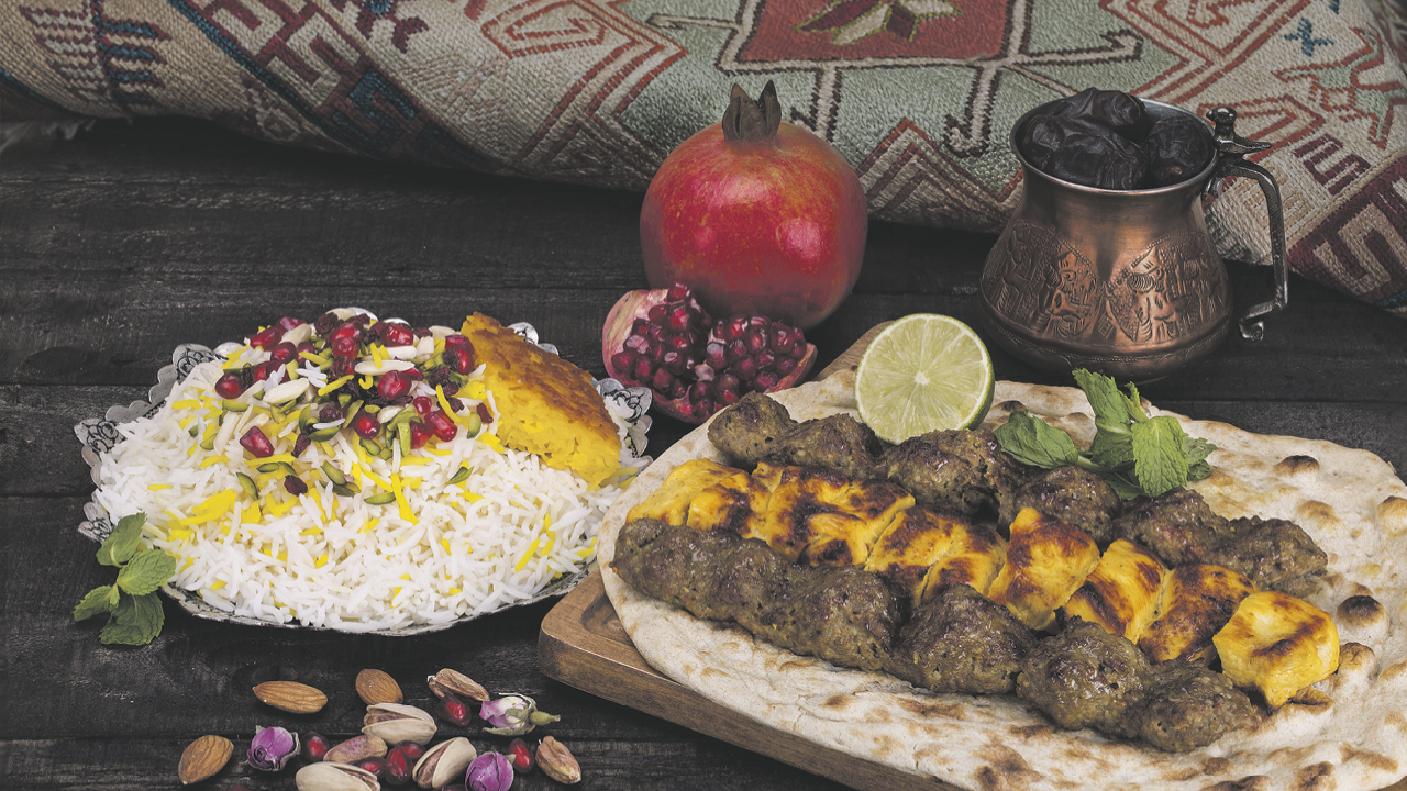 ein persisches Gericht: gebratener Reis mit Berberitzen und Granatapfelkernen, eine Köfte auf einem Fladenbrot mit gegrillten Kartoffeln, Pistazien, ein Granatapfel und eine orientalisches Gefäß mit Pflaumen
