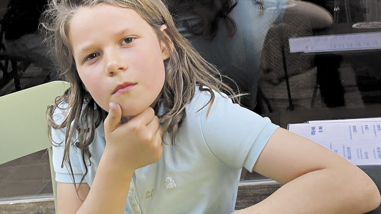 Portrait eines Mädchens, das sitzt und sich mit der Hand das Kinn hält; schaut fragend in die Kamera