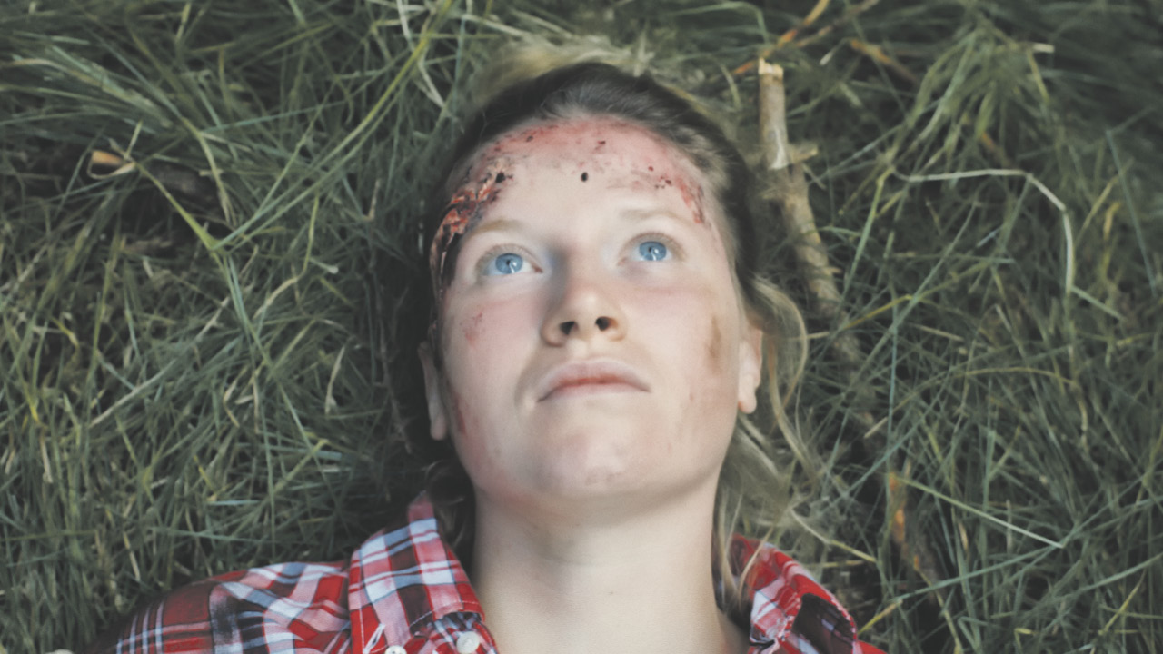 Kopf einer jungen Frau, die mit offenen Augen auf einer Wiese liegt, auf der Stirn Schürfwunden
