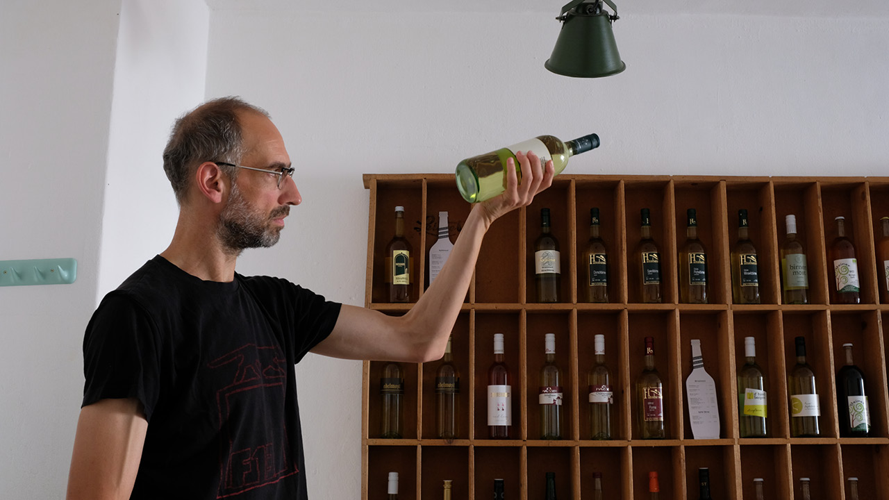 ein Mann mit Brille hält eine Flasche Wein hoch, hinter ihm Regale mit Wein