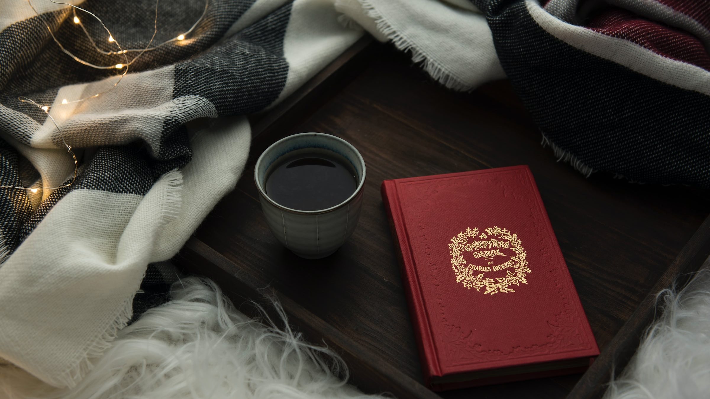 Stillleben mit rot gebundenem Buch und einer Tasse Tee auf einem Holztablett, drumherum eine warme Wolldecke und eine Lichterkette